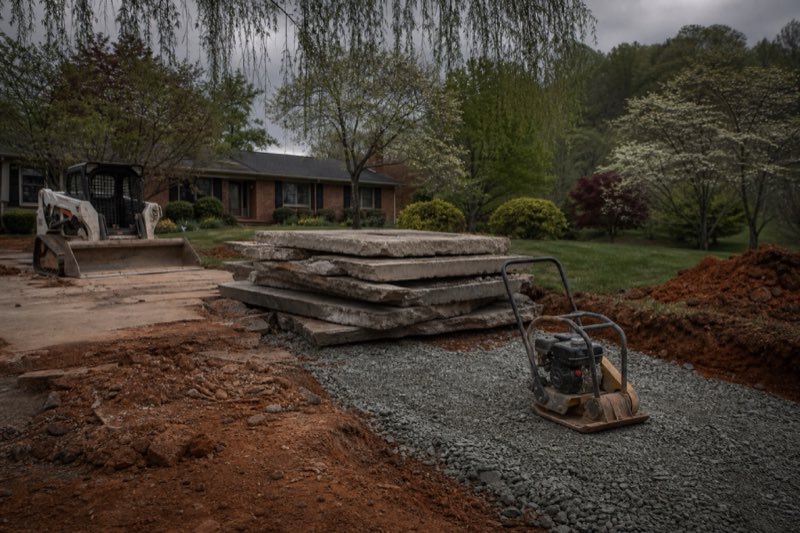 Skid steer loading broken concrete slab pieces into a dumpster during a driveway removal, exposed red clay subgrade in the foreground